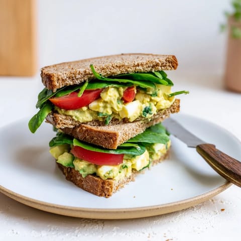 A close-up view shows the vibrant green avocado egg salad sandwich on toasted bread, garnished with chives and lemon zest.  